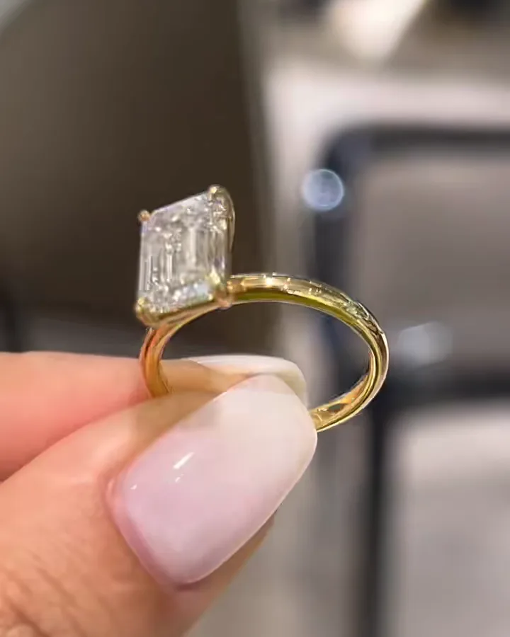 An emerald-cut white stone in claws on a slim gold band, held between two fingers
