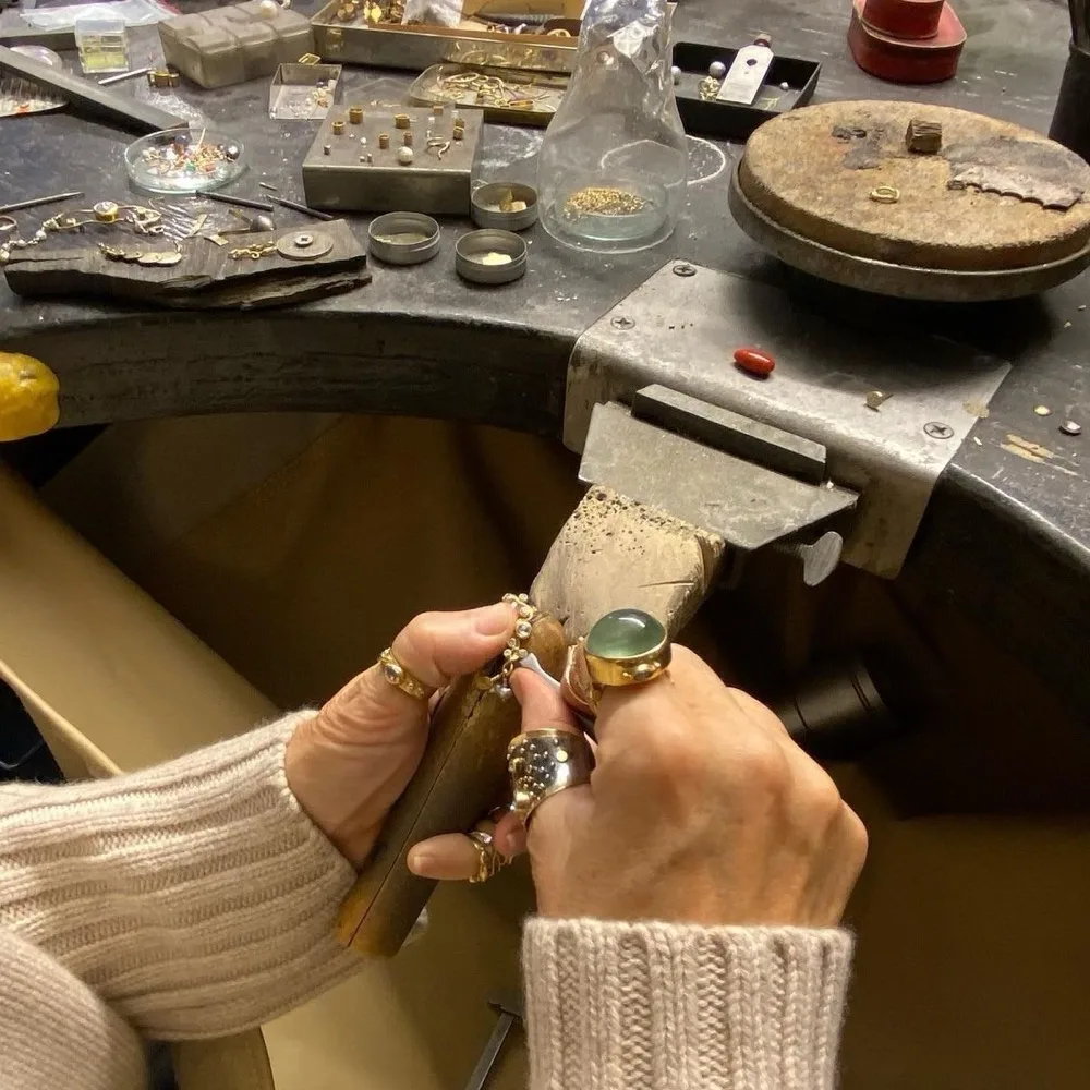 A jeweller's hands at a bench with a vice, wooden peg, jewellery parts and tools.