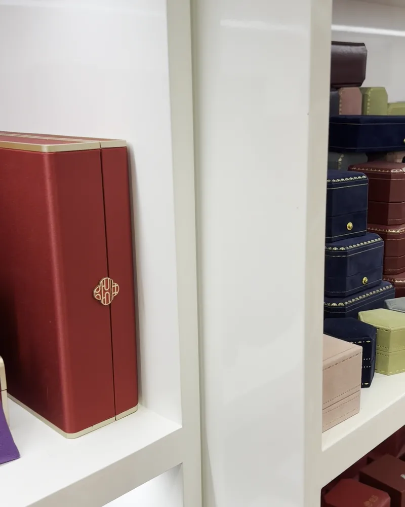 A red jewellery box with a gold clasp on a white shelf, with other boxes beside it
